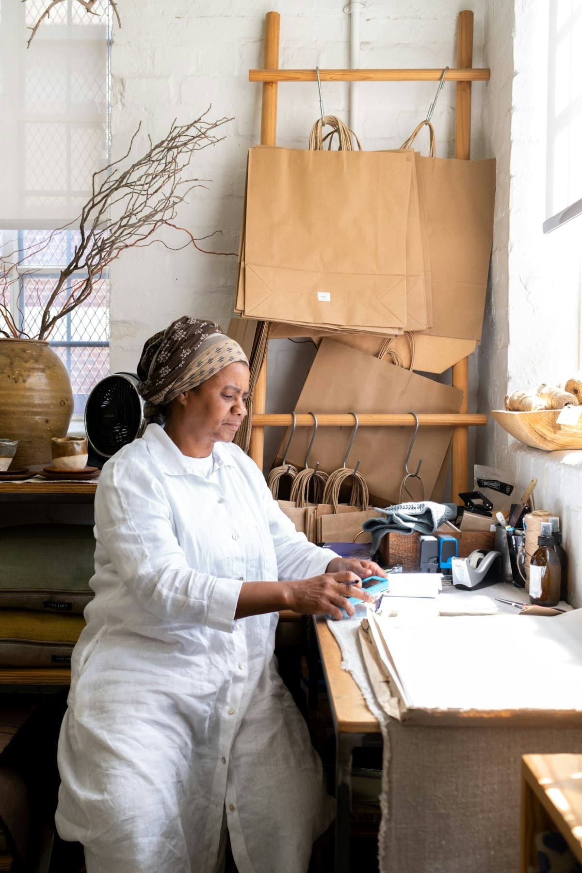 A maker at her workbench in a bright studio, paper shopping bags hanging on the wall behind her